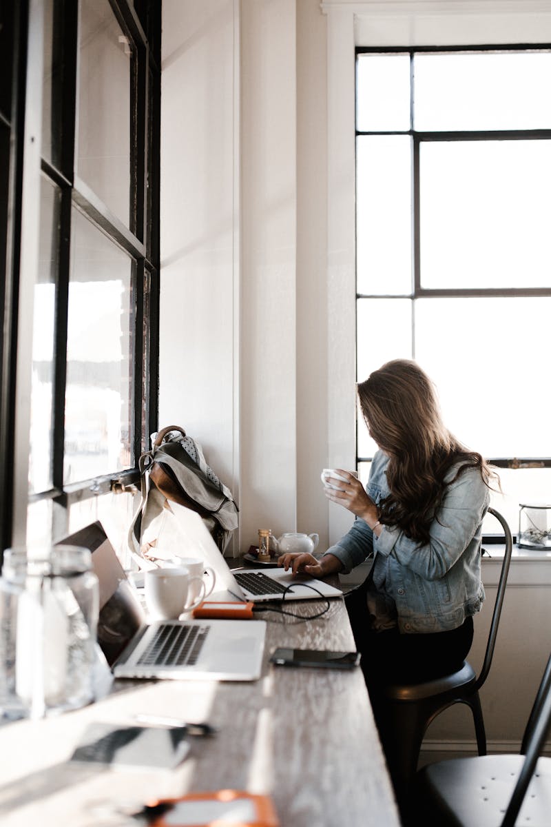 A young woman working on her laptop, sipping coffee in a bright café setting.