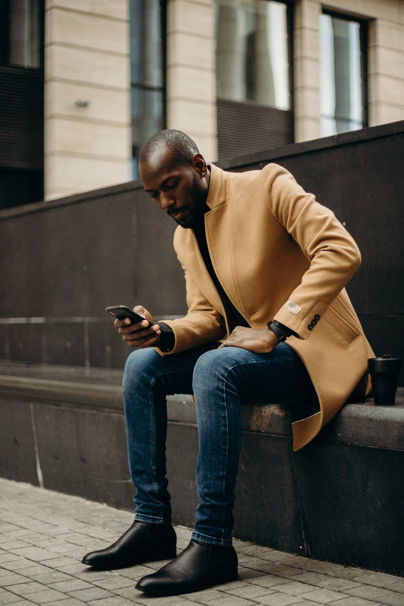 Fashionable man in a trench coat sits outdoors using his smartphone, city setting.