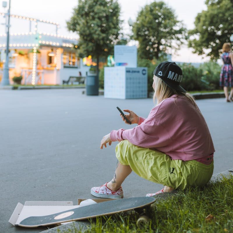 A young woman in casual attire sits on pavement using her smartphone next to a skateboard, enjoying the outdoors.